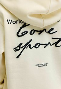 Beige hættetrøje med sort tekst, der lyder "World love sports" i stor skrift og "CORE SPORTSWEAR ASSOCIATION" i små bogstaver.
