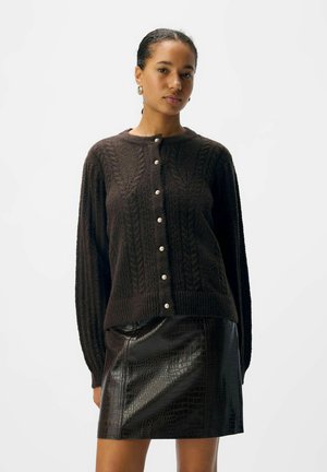 Femme portant un cardigan en tricot à cable marron foncé avec des boutons en perles et une mini jupe en cuir texturé noir sur un fond uni.