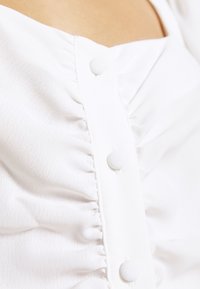 Bílý top z látky s volánkovou texturou a knoflíky potaženými látkou vpředu, vytvářející jemný a decentní design.