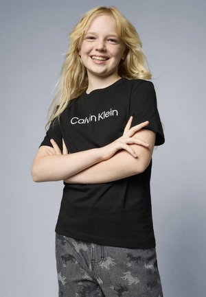 Zwarte katoenen t-shirt met op de voorkant een witte "Calvin Klein" logo, gecombineerd met grijs camouflagepatroon broek.