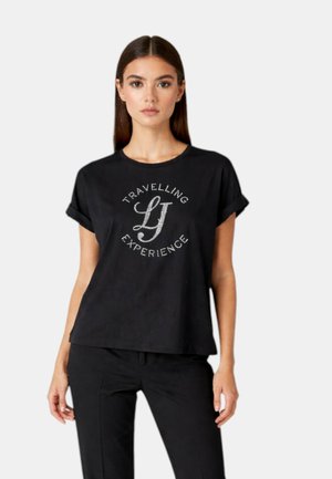 Donna che indossa una T-shirt nera con maniche corte arrotolate e la scritta "Travelling LJ Experience", abbinata a pantaloni neri, in piedi su sfondo bianco.