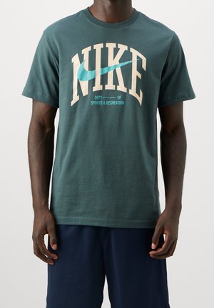 Uomo che indossa una maglietta Nike verde scuro con grande scritta crema "NIKE" e swoosh blu, abbinata a pantaloncini blu scuro, in piedi su sfondo bianco.