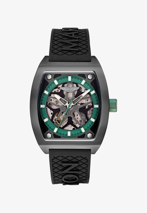 Juodas ir žalias „Aston Martin“ laikrodis su atidarytais svirtimis, stačiakampiu metaliniu korpusu ir tekstūruotu juodu guminiu dirželiu, ant kurio yra įspaustas užrašas "MAR".