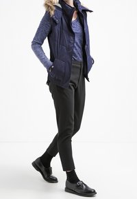 Gilet trapuntato blu navy con rifiniture in pelliccia sintetica, camicia a maniche lunghe blu, pantaloni neri su misura e scarpe nere con fibbie e calze a trama.