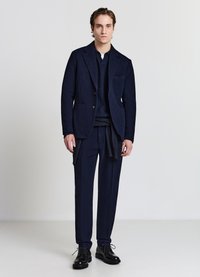 Marineblauer Wollblazer mit Kerbkragen und zwei vorderen Taschen, kombiniert mit passenden Hosen. Schwarze Schnürschuhe vervollständigen das Outfit.