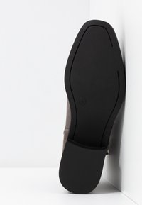 Semelle d'une chaussure habillée à semelle noire appuyée contre un coin blanc, montrant une texture antidérapante et un talon léger.