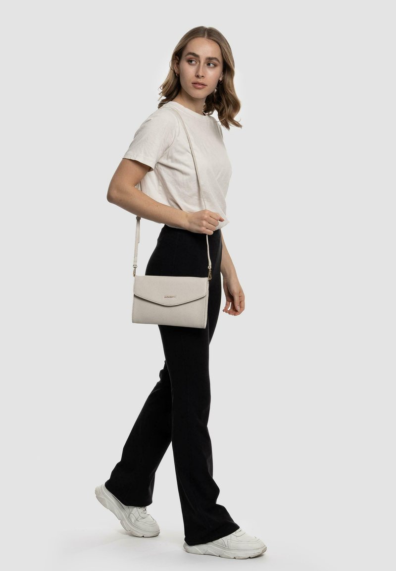 Heller beiger Crossbody-Bag mit strukturiertem Oberflächen, Klappenverschluss und verstellbarem Träger. Getragen von einer Person in einem neutralen Outfit vor einem grauen Hintergrund.
