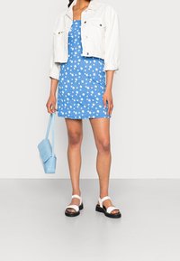 Niebieska sukienka w biały wzór stokrotek, biała krótka jeansowa kurtka, jasnoniebieska torebka oraz białe sandały z czarnymi podeszwami.