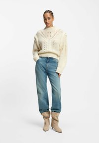 Cream cable-knit sweater with a high neck, featuring a diamond pattern and open stitches, paired with light blue wide-leg jeans and beige heeled boots.
