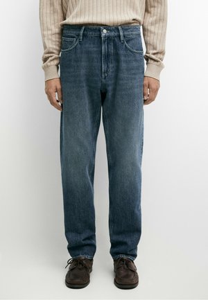 Mand iført straight-fit falmede blå jeans, beige ribbet sweater og brune lædersko, stående mod en ensfarvet baggrund.
