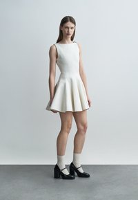 Jonge vrouw staat en draagt een witte mouwloze jurk met een textuur en een uitlopende rok, gecombineerd met witte sokken en zwarte Mary Jane schoenen met blokhakken.