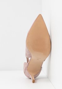 Chaussure à talon haut en daim beige à bout pointu reposant sur le côté contre un mur blanc d'angle, semelle visible affichant la taille 37.