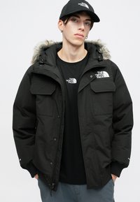 Tânărul îmbrăcat cu o jachetă neagră North Face cu glugă din blană, o ștampilă neagră North Face asortată și o pălărie neagră, stă cu mâinile în buzunare.