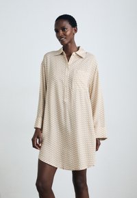 Langärmliges Hemdkleid in hellbeige mit geometrischem Muster, Kragenhals, Fronttasche und einem lässigen Schnitt. Weiche Stoffstruktur.