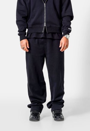 Personne portant un sweat zippé noir, un pantalon noir ample, des baskets noires, une montre argentée et des bagues, debout sur un fond blanc.