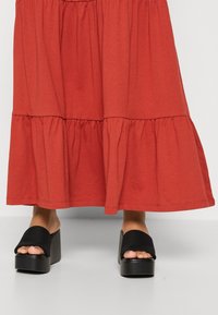 Mules à plateforme noirs avec une large sangle en tissu ; associés à une jupe longue jusqu'aux genoux, fluide, à volants, de couleur rouille faite d'un matériau doux.