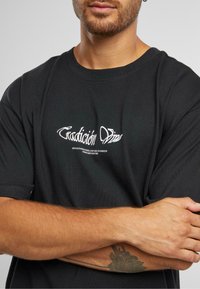 Černé bavlněné tričko se stylizovaným bílým textem "Tradición" a dalším textem níže. Krátké rukávy, design s kulatým výstřihem.