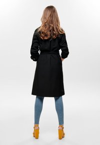 Abrigo negro de manga larga con cinturón en la cintura y puños abotonados, combinado con jeans ajustados de color azul claro y tacones amarillos, visto desde atrás.