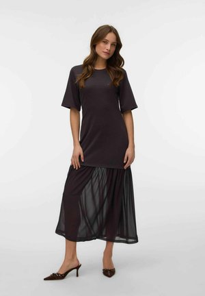 Femme portant une robe courte à manches courtes foncée avec une jupe inférieure transparente et des talons pointus foncés sur un fond clair uni.