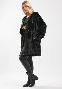 Cappotto lungo in pelliccia ecologica nera con cappuccio, chiusura a zip, due tasche laterali e maniche a coste, abbinato a pantaloni in pelle nera e stivaletti con tacco.