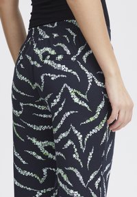 Tmavě modré legíny s květinovým vzorem, elastickým pasem a boční kapsou, které na sobě mají bílé a zelené květinové vzory vlnkového tvaru.