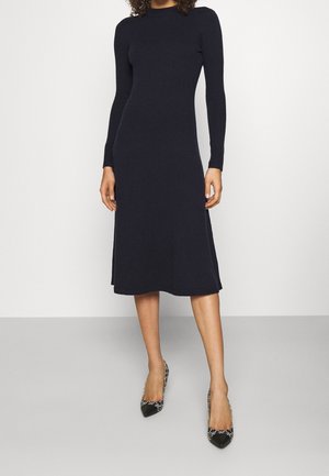 Vrouw draagt een nauwsluitende, lange mouwen, zwarte midi-jurk met zwart gemêleerde puntige hakken, staand tegen een effen lichte achtergrond.