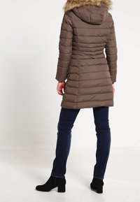 Brązowa kurtka puchowa z kapturem wykończonym futrem, z pionowym pikowaniem, w zestawieniu z ciemnoniebieskimi dżinsami i czarnymi botkami za kostkę.