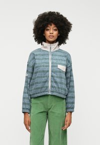 Ελαφρύ μπουφάν με φερμουάρ σε τόνους teal με υφή ριγέ. Διαθέτει ψηλό γιακά, ελαστικούς καρπούς και μικρή τσέπη στο στήθος.