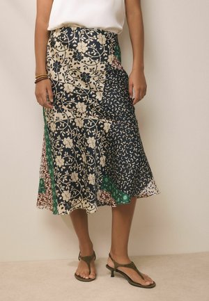 Kobieta nosząca midi spódnicę z mieszanymi kwiatowymi i geometrycznymi wzorami w granacie, kremie, zieleni i burgundzie, w połączeniu z oliwkowo zielonymi sandałami.