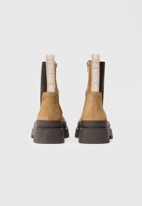 Botas de tobillo de ante en color marrón claro con una suela negra gruesa, que presentan un panel lateral elástico negro en contraste y una pestaña con logotipo en la parte trasera.
