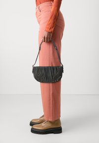 Geantă din denim închis cu capse aurii, design plisat și un mâner scurt, ținută de o persoană care poartă pantaloni roz și bocanci maro cu șireturi.