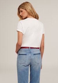 Camisa de manga corta blanca con puños enrollados, combinada con jeans de mezclilla azul claro y un cinturón rojo. Presenta un diseño estándar de bolsillo trasero.