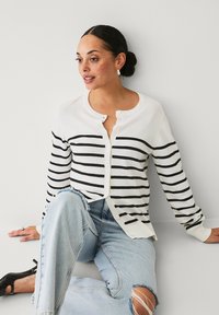 Cardigan bianco con strisce nere, chiusura a bottoni e polsini a coste. Abbinato a jeans azzurri strappati e tacchi neri.