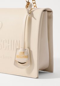 Beige kožená kabelka s vyraženým textem "MOSCHINO", zlatým řetízkovým popruhem a štítkem s nápisem "LOVE MOSCHINO" v zlatém písmu. Hladký povrch.
