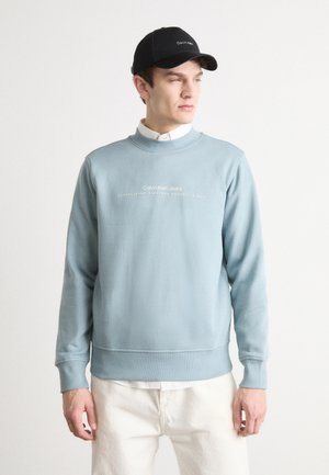 Hellblaues Sweatshirt mit Rundhalsausschnitt und langen Ärmeln, ausgestattet mit weißem Logotext. Kombiniert mit einem weißen Polohemd und cremefarbenen Hosen. Schwarze Kappe.