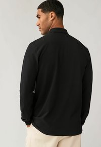 Zwarte lange mouw polo met een zachte textuur. Heeft een kraag en subtiele stikdetails, gedragen met lichtgekleurde broek.