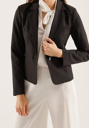 Vrouw draagt een zwart, getailleerd colbert over een witte blouse met een gestrikte halsstrik en witte hoge taillebroeken.