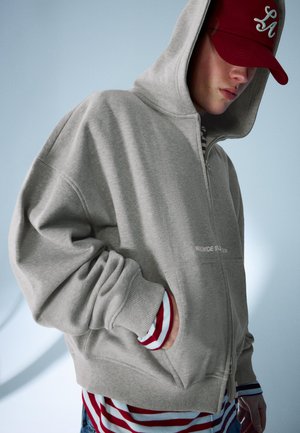 Jeune personne portant un sweat à capuche gris zippé par-dessus un t-shirt rayé rouge et blanc ainsi qu'une casquette rouge, main dans la poche, regard dirigé vers le bas.