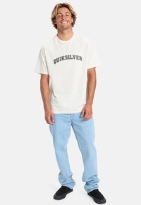 Creme T-shirt med "Quiksilver" tekst i en kontrastfarvet mørk skrift, parret med lyseblå jeans og sorte casual sko. Enkel, afslappet pasform.