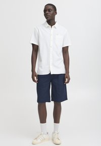 Biela košeľa s krátkym rukávom na gombíky s vreckom, v kombinácii s námorníckymi modrými šortkami a svetlými teniskami. Jednoduchý dizajn, textúrovaná tkanina a neutrálne farby.