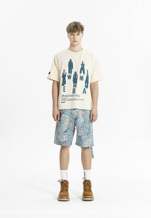 Joven de pie, vistiendo una camiseta gráfica beige con siluetas y texto, pantalones cortos azules con estampado, calcetines blancos y botas marrones, fondo blanco liso.