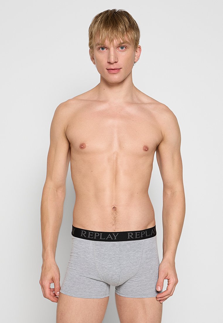 Junger Mann steht mit entspannten Armen, trägt graue Boxershorts mit schwarzem "REPLAY"-Bund, vor einem schlichten, hellen Hintergrund.