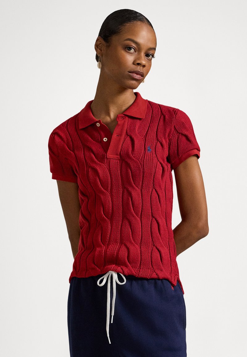 Rdeča pletena polo majica z vzorcem pletenja kabla, kontrastnim ovratnikom, vezenim logotipom in kratkimi rokavi. Stilizirana z mornarsko modrimi hlačami z zatezno vrvico na pasu.