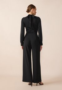 Blouse noire à manches longues avec un nœud à l'arrière du cou, associée à un pantalon noir à jambes larges et des sandales noires à talons. Texture de tissu lisse.