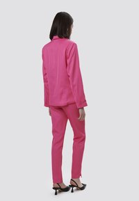 Completo rosa aderente con giacca su misura e pantaloni abbinati. Materiale morbido, colletto elegante e dettagli discreti sulla cucitura posteriore, abbinato a tacchi neri.