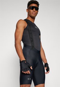 Svart sykkel bodysuit med mesh-detaljer, med justerbare stropper. Svart fingerløse hansker fremhever grep. Reflekterende logo aksenter er synlige.