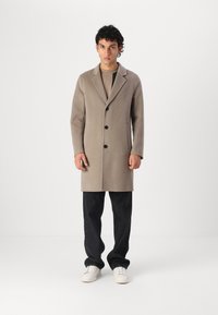 Cappotto in lana beige con colletto a rotondo, tre bottoni neri e tasche laterali, abbinato a pantaloni neri e sneakers bianche.