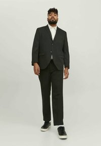Black suit with a single-button blazer and two front pockets, paired with matching trousers. Worn with a white shirt and black sneakers.
