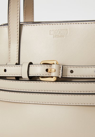 Sac à main en cuir beige avec une boucle dorée et des détails de couture. Le logo en relief "Moschino Milano" est présent sur la surface.