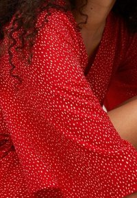 Femme aux cheveux bouclés portant une chemise à manches longues rouge avec des taches blanches, avec une partie de son cou et de son bras visible.